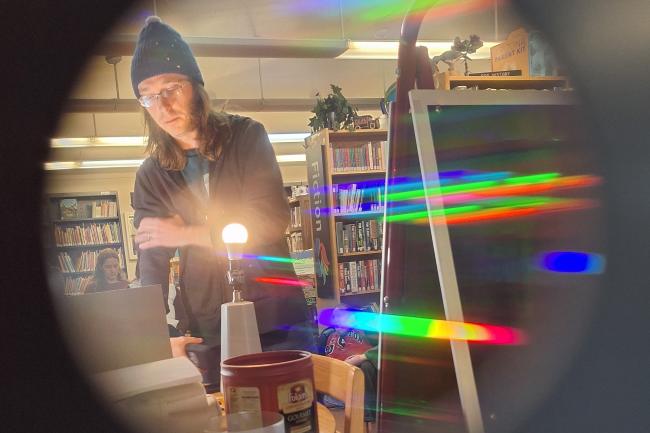 Camera view through a round hole. Reflections of light off of lightbulbs show a spectrum of color with man in beanie in the background. 