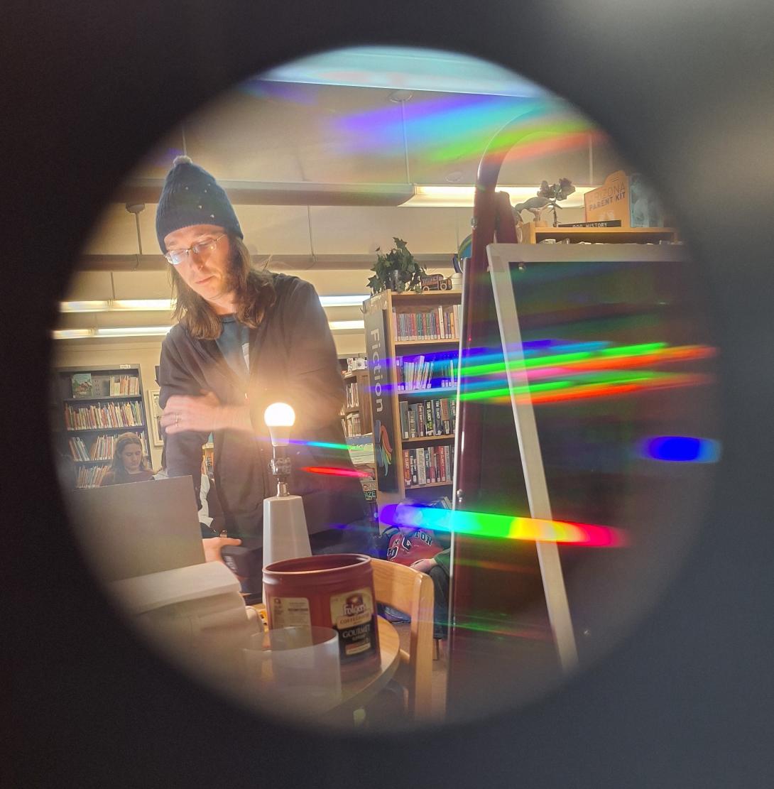 Camera view through a round hole. Reflections of light off of lightbulbs show a spectrum of color with man in beanie in the background. 